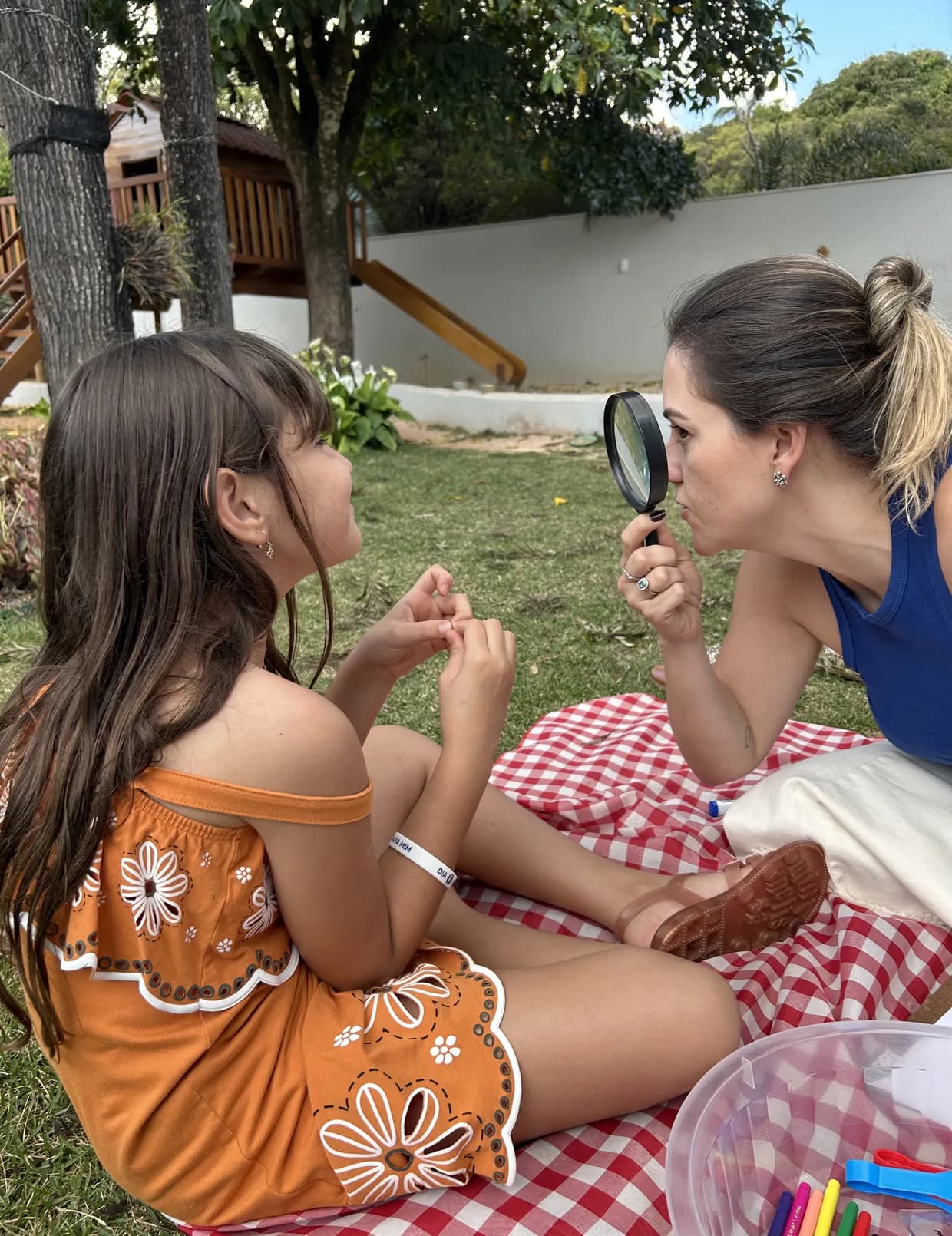 Educadora e criança sentadas numa toalha xadrez no gramado, observando algo de perto com uma lupa